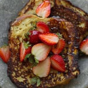 Two slices of golden-brown vegan French toast topped with fresh strawberries and a drizzle of syrup, served on a gray plate.